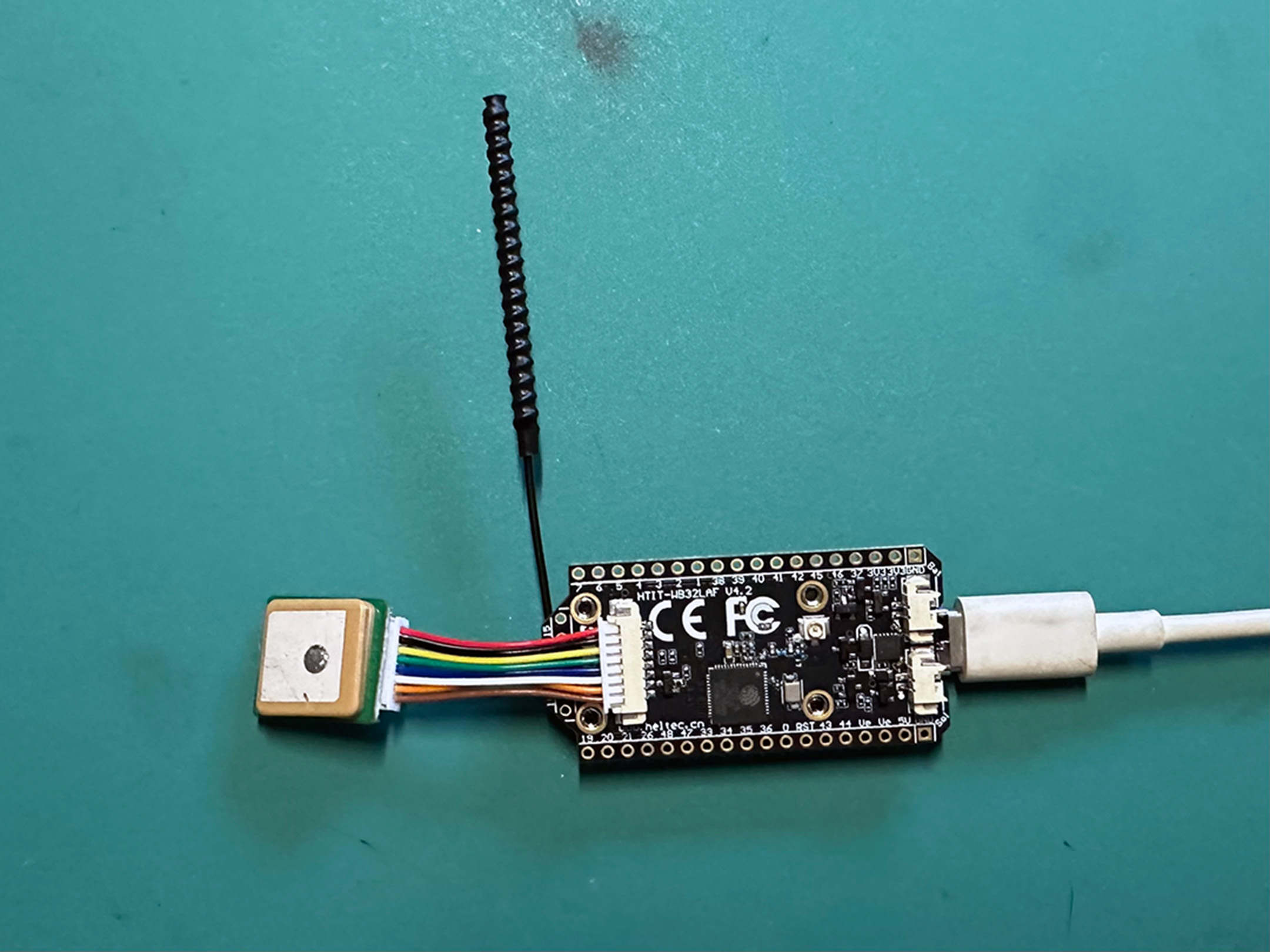 Heltec HTIT-WB32LAF v4.2 board with GNSS patch antenna, spring LoRa antenna, and USB-C cable on a teal work mat.